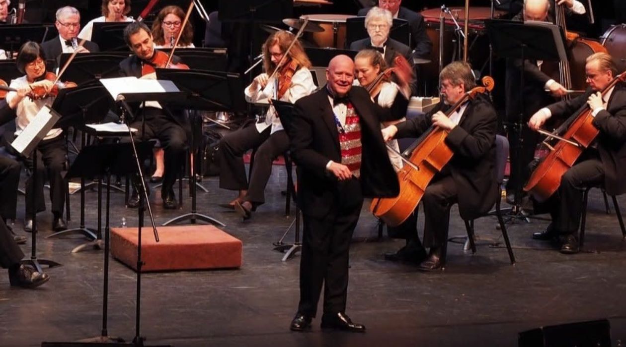 The conductor of Orchestra New England, dressed in a black tuxedo with a glittery vest that resembles the American flag, is surrounded by orchestra members playing various string instruments during a concert.